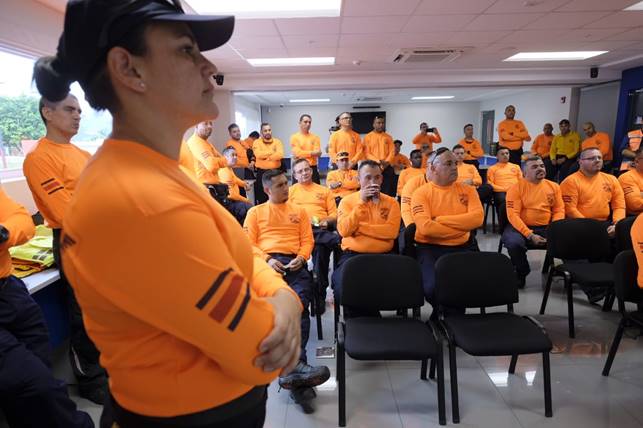 Se aprecian rescatistas con su uniforme anaranjado, unos de pie y otros sentados, en un aula recibiendo una charla.