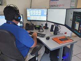 Fotografía de un funcionario de la CNE, de camisa azul, sentado frente a un escritorio viendo tres pantallas de una computadora y escribiendo con el teclado. Tiene unos audífonos puestos.